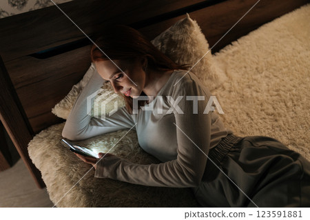 Top view of pretty young woman relaxing in bed, scrolling smartphone with soft bedroom lighting, enjoying late night digital connection and personal downtime. Concept of mobile addiction and insomnia. Top view of pretty young woman relaxing in bed, scrolling smartphone with soft bedroom lighting, enjoying late night digital connection and personal downtime. Concept of mobile addiction and insomnia. 123591881