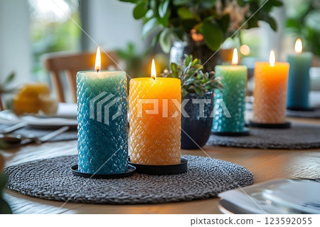 A close-up of lit textured candles in blue and orange hues on a dining table with a blurred background of greenery 123592055