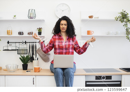 African woman seated in modern kitchen using laptop, practicing meditation, tranquil atmosphere. White minimalist kitchen style, indoor plants decorate space. 123599647