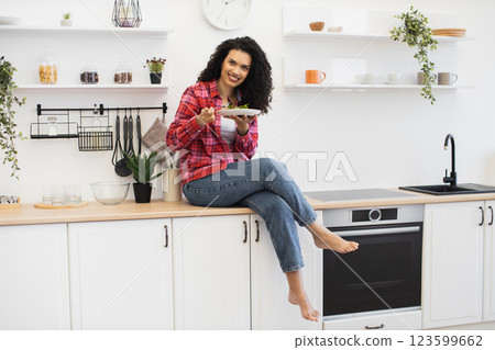 Joyful young African woman in a casual kitchen setting sitting on counter holding plate. Bright and modern interior with a cozy vibe showcasing home lifestyle and meal enjoyment. Joyful young African woman in a casual kitchen setting sitting on counter holding plate. Bright and modern interior with a cozy vibe showcasing home lifestyle and meal enjoyment. 123599662