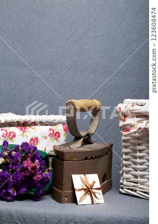 Old iron, heated by hot coals. Located on gray fabric. Nearby are wicker baskets, a bouquet of dried flowers and a greeting card. 123608474