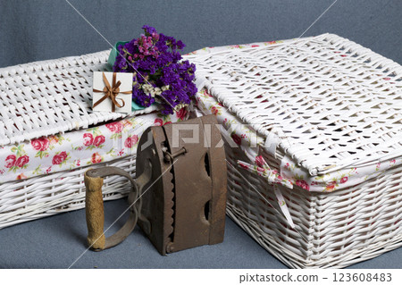 Old iron, heated by hot coals. Located on gray fabric. Nearby are wicker baskets, a bouquet of dried flowers and a greeting card. 123608483