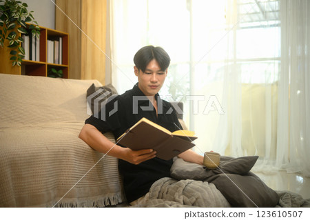 Young man holding a cup of coffee and reading book in a cozy living room 123610507