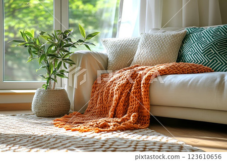 modern cozy living room with a soft beige sofa, textured pillows, an orange knitted blanket, and a potted plant 123610656