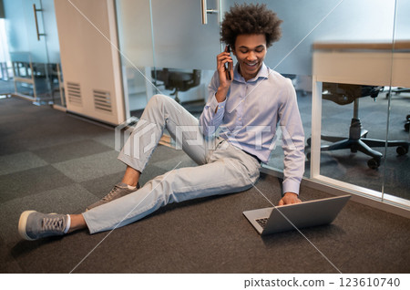 Male student freelancer using computer for working on office floor and talking on smartphone Male student freelancer using computer for working on office floor and talking on smartphone 123610740