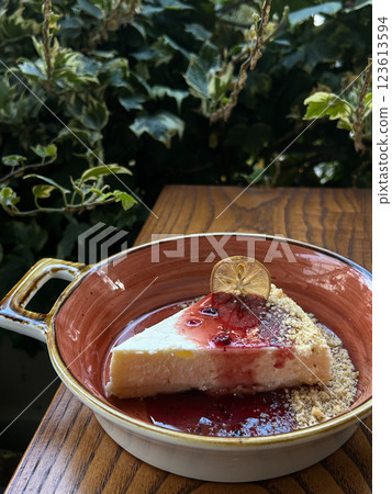 Cheesecake slice with berry sauce and dried lemon garnish in a ceramic dish on a wooden table. Suitable for food photography, restaurant menus, catering, and social media 123613594