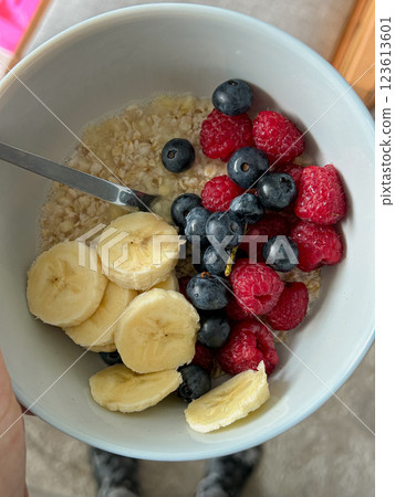 A close-up of a white bowl filled with oatmeal, banana slices, raspberries, and blueberries with a spoon. The image is suitable for food blogging, nutrition, and wellness content 123613601