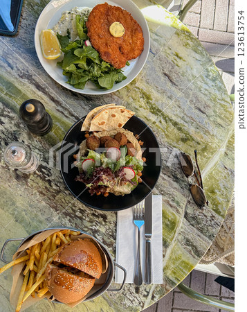 A stylish outdoor dining scene featuring a crispy schnitzel, a falafel plate, and a gourmet burger with fries, set on a marble table under natural light 123613754