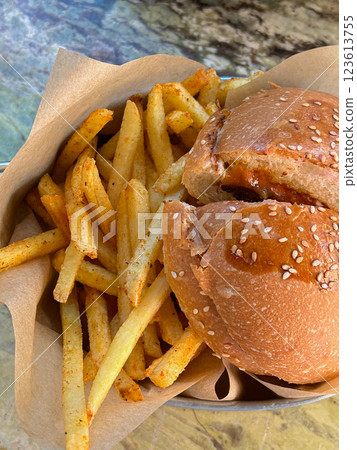 A burger with a sesame bun and a portion of seasoned fries served in a metal tray lined with parchment paper on a marble table. Suitable for food blogs and restaurant marketing 123613755