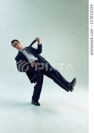 Businessman pulling tie playfully, leaning back, balancing on one leg against white studio background. Concept of business, stress, humor, life balance. 123614284