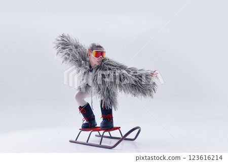 Young woman balancing on sled in oversized shaggy fur coat, ski goggles, and moon boots against white studio background. Young woman balancing on sled in oversized shaggy fur coat, ski goggles, and moon boots against white studio background. 123616214