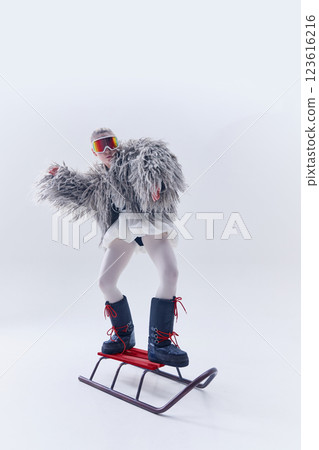 Young woman striking pose on sled in oversized shaggy fur coat, ski goggles, and moon boots against white studio background 123616216