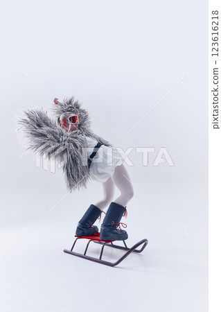 Young woman leaning back on sled in oversized shaggy fur coat, ski goggles, and moon boots against white studio background. Young woman leaning back on sled in oversized shaggy fur coat, ski goggles, and moon boots against white studio background. 123616218