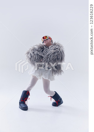 Young woman striking stylish pose in oversized shaggy fur coat, ski goggles, and moon boots against white studio background 123616229