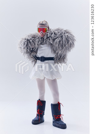 Young woman standing confidently in oversized shaggy fur coat, ski goggles, and moon boots against white studio background 123616230