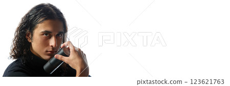 Young man in black turtleneck holding black deodorant, perfume bottle with intense expression against white studio background. 123621763