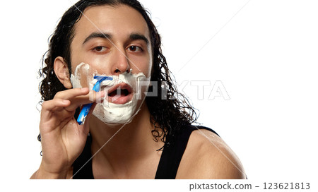 Young man with curly wet hair doing morning routine, shaving face with blue razor, thick foam covering skin against white studio background. 123621813