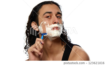 Young man with curly wet hair shaving face with blue razor, thick foam covering skin against white studio background. 123621816