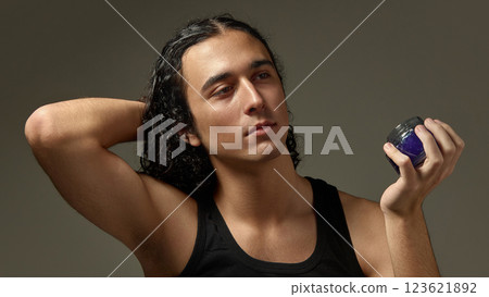 Handsome young guy with flawless skin gently touching curly hair while holding premium hair product against minimalist studio setting. 123621892