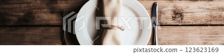 Simple and elegant table setting with a white plate, silver cutlery, and a beige napkin on a rustic wooden surface. Warm natural light enhances the cozy, minimalistic, country-style ambiance. 123623169