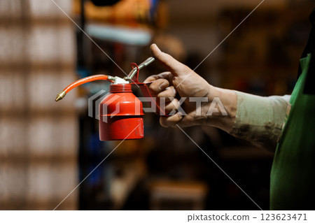 Close-up View of Mechanic's Hand Holding Red Oil Lubricating Can in Workshop Environment 123623471