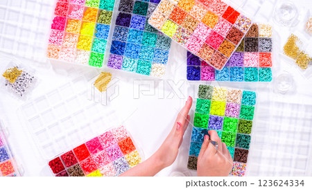 Flat lay. Woman hands gracefully poised over a collection of beads, sorted by color in transparent organizers. The array of beads spans a vibrant spectrum, from deep purples to bright oranges 123624334