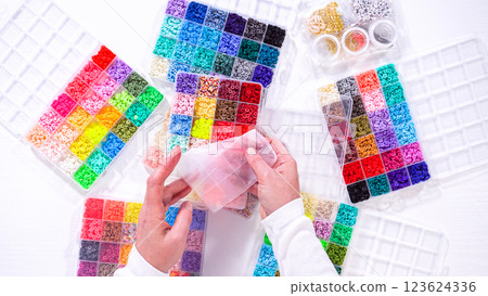 Flat lay. Woman hands gracefully poised over a collection of beads, sorted by color in transparent organizers. The array of beads spans a vibrant spectrum, from deep purples to bright oranges 123624336