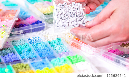 Woman hands gracefully poised over a collection of beads, sorted by color in transparent organizers. The array of beads spans a vibrant spectrum, from deep purples to bright oranges, meticulously Woman hands gracefully poised over a collection of beads, sorted by color in transparent organizers. The array of beads spans a vibrant spectrum, from deep purples to bright oranges, meticulously 123624341
