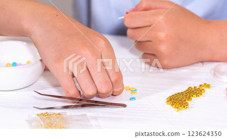 Delicate fingers of a young girl navigate through a treasure trove of bright, multicolored beads, each compartment revealing a new hue to choose from. She immersed in the joyful task of stringing 123624350