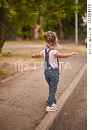 The girl walks along the sidewalk, keeping her balance, arms outstretched. 123624629