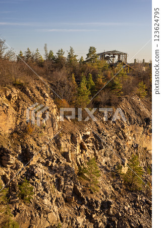 A rocky cliff with sparse trees and dry bushes under warm sunlight. At the top, an abandoned industrial structure stands among autumn foliage, creating a contrast between nature and human-made ruins. 123624795