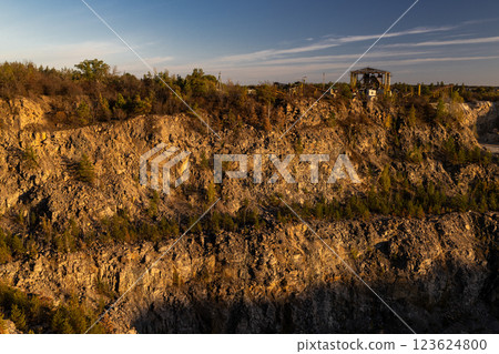 A rocky cliff with sparse trees and dry bushes under warm sunlight. At the top, an abandoned industrial structure stands among autumn foliage, creating a contrast between nature and human-made ruins. A rocky cliff with sparse trees and dry bushes under warm sunlight. At the top, an abandoned industrial structure stands among autumn foliage, creating a contrast between nature and human-made ruins. 123624800