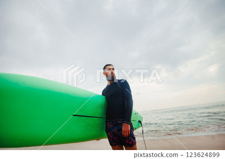 Young handsome male surfer with a surfboard is enjoying a view while walking a sandy beach at sea. Summer, vacation, ocean Young handsome male surfer with a surfboard is enjoying a view while walking a sandy beach at sea. Summer, vacation, ocean 123624899