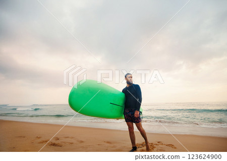 Young bearded man with surfboard standing near a beach. Man with surfing board outdoors on a summer day Young bearded man with surfboard standing near a beach. Man with surfing board outdoors on a summer day 123624900
