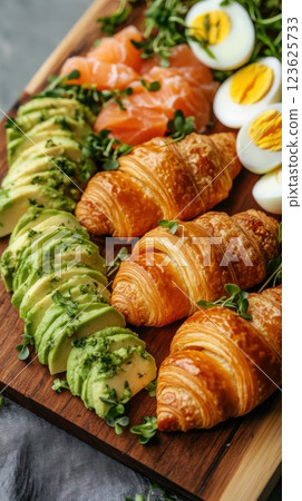 Low-angle shot of a wooden board with golden croissants, fresh avocado, boiled eggs, salmon, and microgreens. Vibrant, dynamic presentation with natural light and rustic warmth 123625733