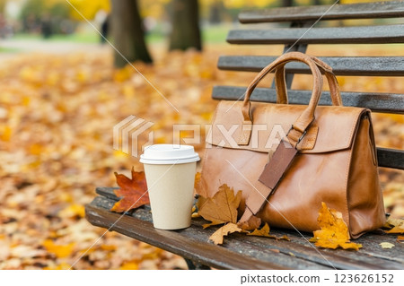 A leather handbag rests on a park bench surrounded by colorful autumn leaves and a warm coffee cup 123626152