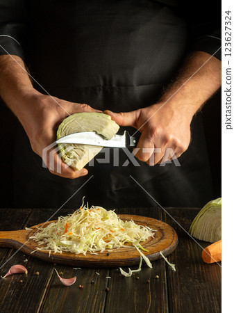 A chef skillfully slices a fresh cabbage in a rustic kitchen. Shredded cabbage and vegetables lie on a wooden board, showcasing expert culinary skills and fresh ingredients 123627324