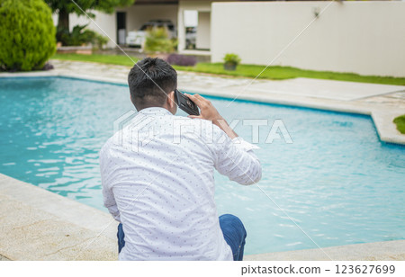 Handsome man sitting calling on the phone near the swimming pool, Young man sitting calling on the phone by a swimming pool, Guy sitting talking on the cell phone near the edge of the swimming pool 123627699