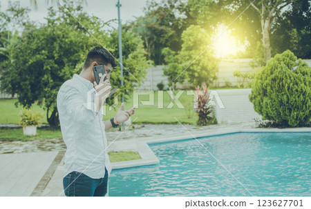 Attractive guy standing talking on the phone near the pool edge. Handsome man calling on the phone by a swimming pool, Young man standing calling on the phone by a swimming pool Attractive guy standing talking on the phone near the pool edge. Handsome man calling on the phone by a swimming pool, Young man standing calling on the phone by a swimming pool 123627701