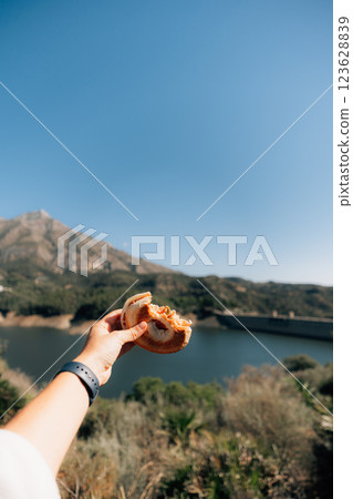 Hand holding bagel with mountain and lake view under clear blue sky 123628839