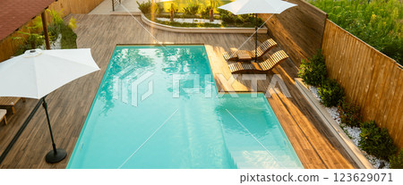 Modern swimming pool with turquoise water, wooden deck, umbrellas, and lounge chairs in a tranquil backyard Modern swimming pool with turquoise water, wooden deck, umbrellas, and lounge chairs in a tranquil backyard 123629071
