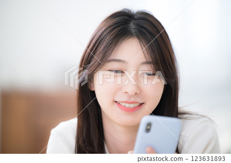 特寫：一位年輕女子麵朝下躺在房間裡，看著自己的智慧型手機 123631893