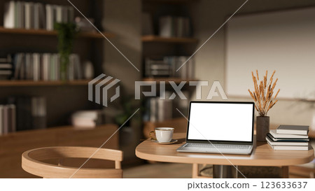 The white screen laptop place between a cup and books on a round wooden table in the dim light room 123633637