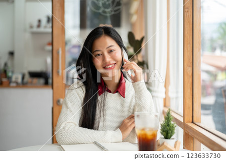 An asian woman is smiling during phone call while looking straight into the camera. sitting in cafe. An asian woman is smiling during phone call while looking straight into the camera. sitting in cafe. 123633730