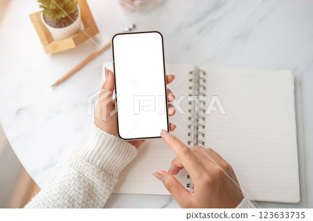 The hand holding blank screen smartphone over notebook with a finger pointing at the screen on table 123633735