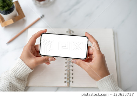 Close up of the pair of hand holding smartphone horizontally over an open notebook on marble table. 123633737