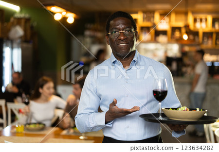 African man of public catering cafe carries order to client, ready to serve guest of cafe establishment. African man of public catering cafe carries order to client, ready to serve guest of cafe establishment. 123634282