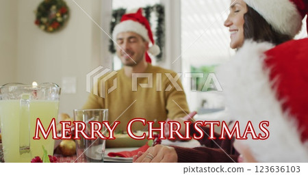Image of merry christmas text over happy caucasian family in santa hats saying grace at dinner 123636103
