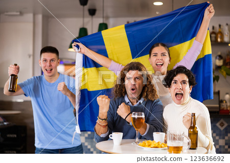 Company of excited young adult sports fans waving flag of Sweden and supporting national team with beer in bar Company of excited young adult sports fans waving flag of Sweden and supporting national team with beer in bar 123636692