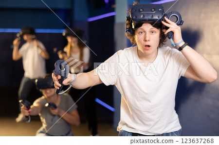 Emotional young guy in VR headset with joysticks playing with friends Emotional young guy in VR headset with joysticks playing with friends 123637268